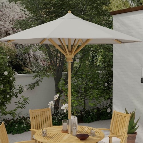 17540F Somerset 8 foot Round teak Umbrella extended on round teak dining set with 2 glasses and white flowers in vase a candle and bowl on tabletop with landscape plants and house in background