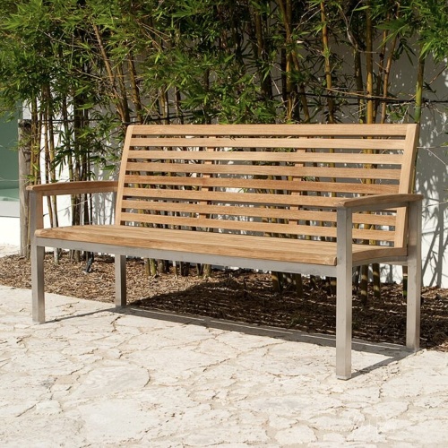 23200 Vogue 5 foot Teak and Stainless Steel Bench angled on concrete patio with shrubs and glass sliding doors in background