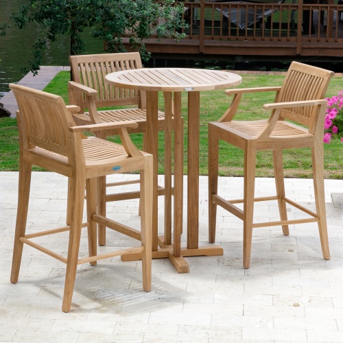 70504 Laguna 4 piece teak Pub Set of 3 barstools with armrests and Round 30 inch diameter Table on stone patio with flowering plant grass area screened boathouse and lake background