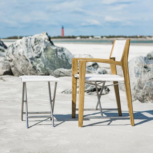 70769 odyssey teak and stainless steel folding chair and ottoman set on sand with boat peer in background