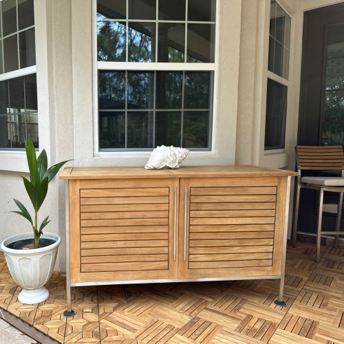 28226 Vogue Teak and Stainless Steel Beverage Console next to a potted plant on teak tiled outdoor patio with windows of a house in the background
