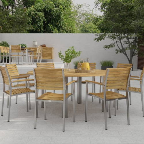70111 Vogue teak and stainless steel 9 piece72 inch round Dining Set with a vase and a teak tray with a pitcher and 2 glasses in angled side view on stone patio with wall and plants in back