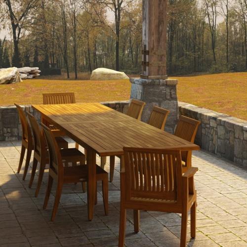 70123 Laguna 9 pc Teak Dining Set of Laguna 11 ft Rectangle Dining Table and 2 Dining Armchairs and 6 Side Chairs in angled end view on stone patio with grass and woods in the background