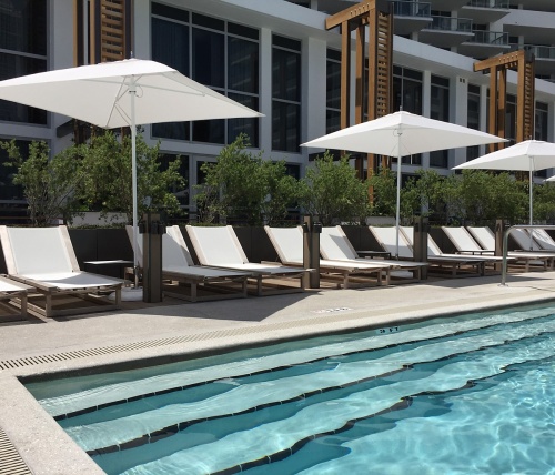 70780 Maya teak Sling Lounger showing 11 on pool deck front facing pool with three rectangular umbrellas with landscape plants trees and hotel in background
