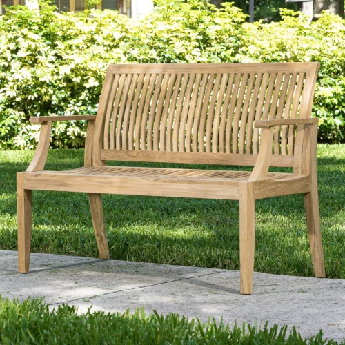 13810 Laguna 4 foot Teak Bench angled on concrete walkway with bushes in background