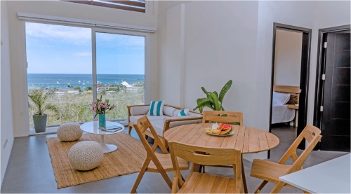70519 Surf 5 piece teak round Dining Set with fruit bowl in center and 2 potted plants teak loveseat 2 poofs white coffee table with vase and glass sliding doors and ocean in background