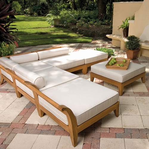 16766DP Aman Dais End Sectional Set side angled view with bolsters and cushions and a teak serving tray with green apples on an ottoman on a paver patio with a grass and tree background