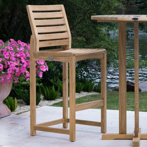  70512 Somerset barstool angled next to teak round table with flowering plants and shrubs in background