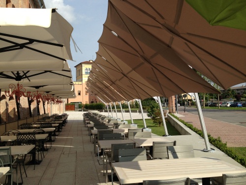 SP25100SET Spectra Solo Umbrella and Paver Base at an outdoor dining area next to paved sidewalk with grass trees buildings and parking lot in background