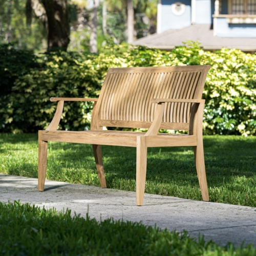 13810 Laguna 4 foot Teak Bench angled side view on concrete walkway with bushes and house in background